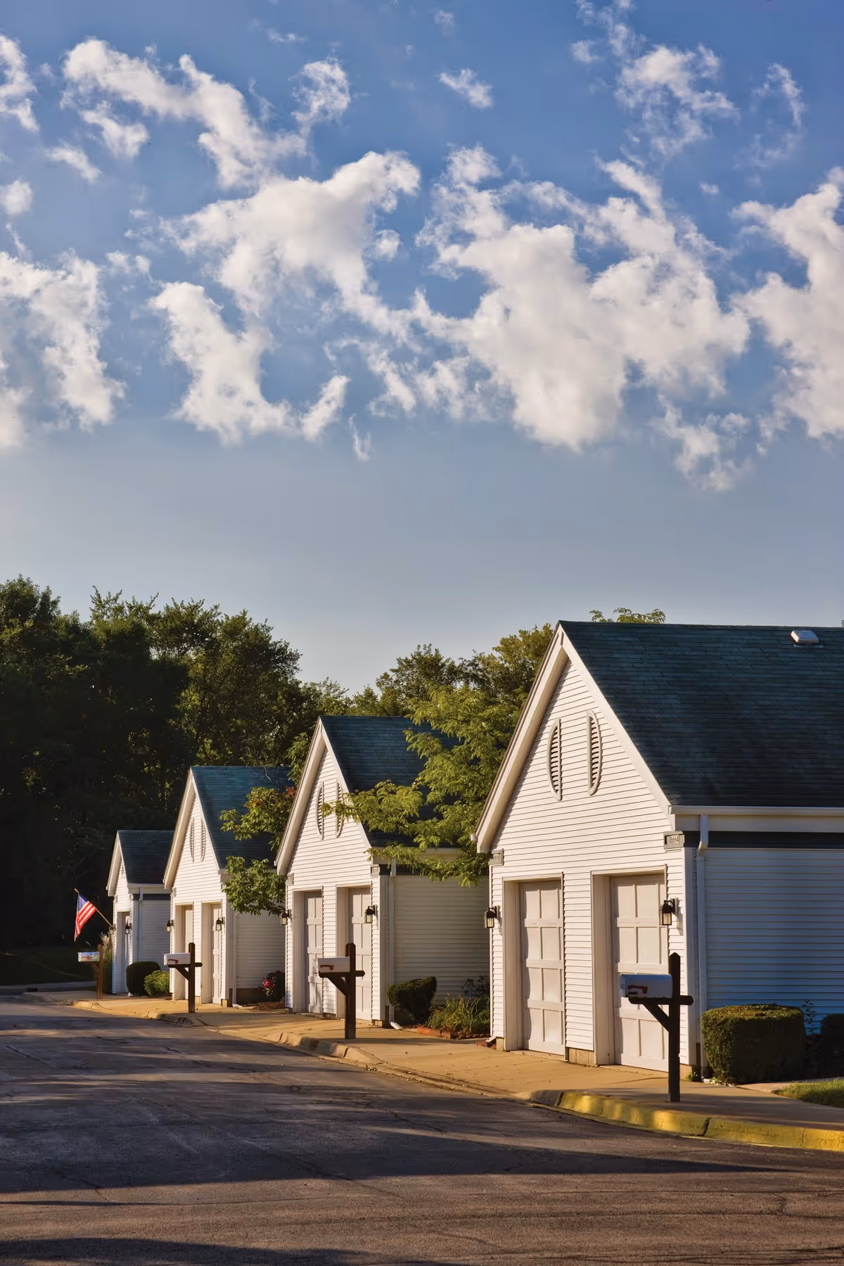 A row of white garage-style cottages with mailboxes along a paved road under a blue sky with scattered clouds.