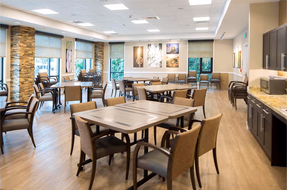 Spacious senior living dining room with multiple tables and chairs, large windows, artwork on the walls, and a kitchenette counter.