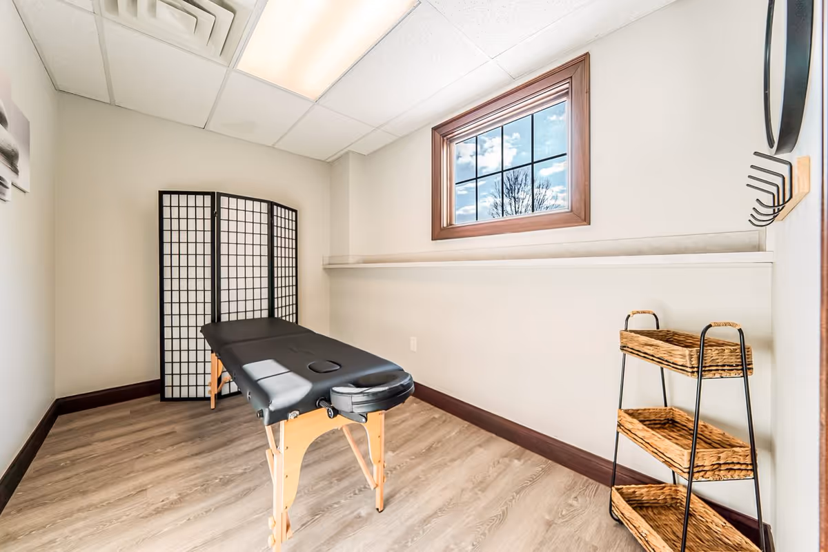 A small, bright room with light-colored walls and wood flooring featuring a black massage table in the center. There is a three-panel folding screen in the corner, a window with a wooden frame letting in natural light, and a three-tier wicker basket shelf on the right side. A wall-mounted rack with hooks is also visible on the right wall.