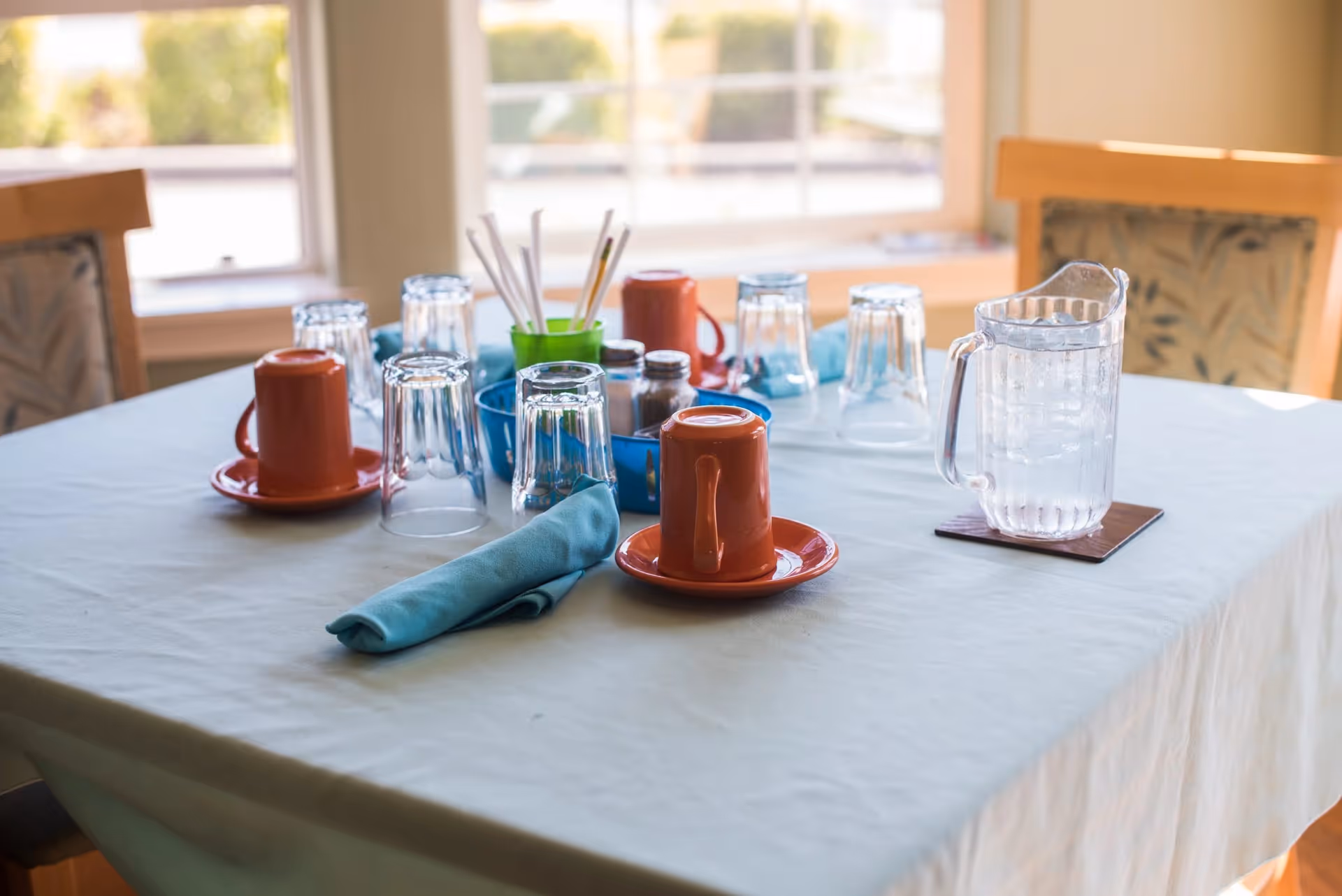 A dining table set with upside-down orange coffee mugs on matching saucers, several upside-down clear drinking glasses, a pitcher of water with ice on a coaster, blue cloth napkins, and a blue tray holding salt and pepper shakers and a green cup with straws, in a room with large windows and wooden chairs with patterned cushions.