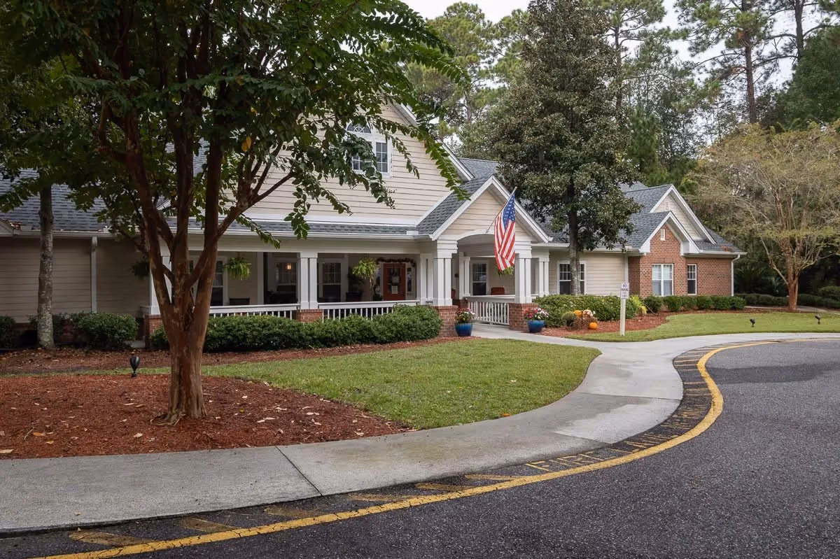 Exterior view of a senior living facility building with a covered porch, white columns, and an American flag. The building is surrounded by well-maintained landscaping including bushes, trees, and flower pots. A curved driveway and sidewalk lead to the entrance.