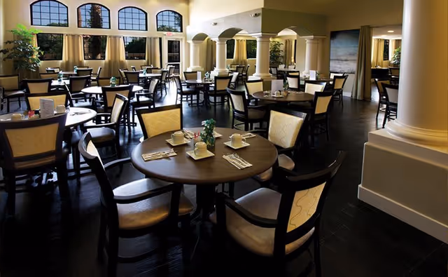 A spacious dining room with multiple round tables set with cups, saucers, napkins, and utensils. The room features large windows with arched tops and beige curtains, cream-colored walls, and decorative columns. The floor is dark wood, and the lighting creates a warm and inviting atmosphere.