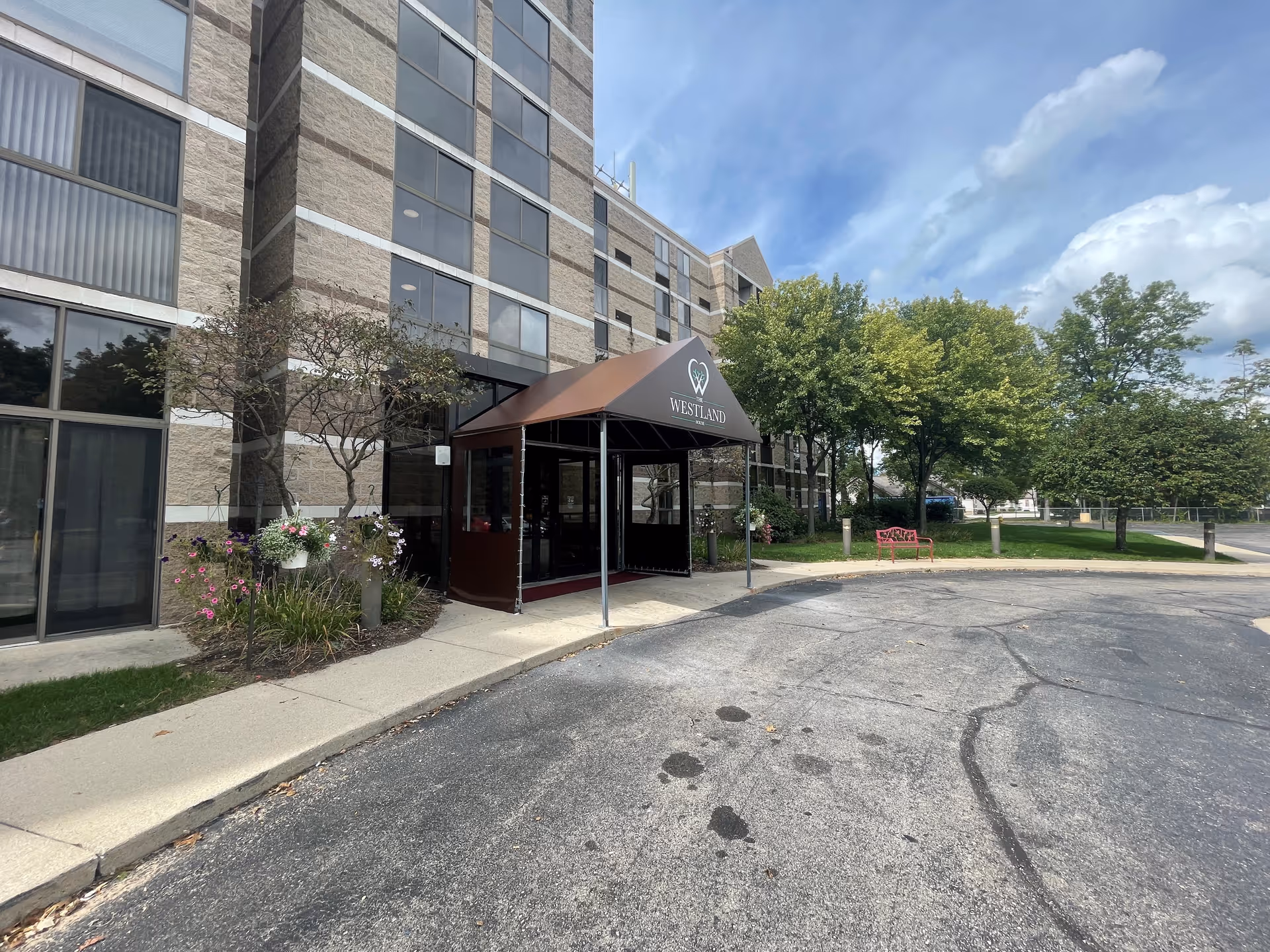 Exterior view of The Westland House building entrance with a brown canopy displaying the facility's name and logo. The building is multi-story with large windows, surrounded by trees, flowers, and a red bench near the sidewalk.