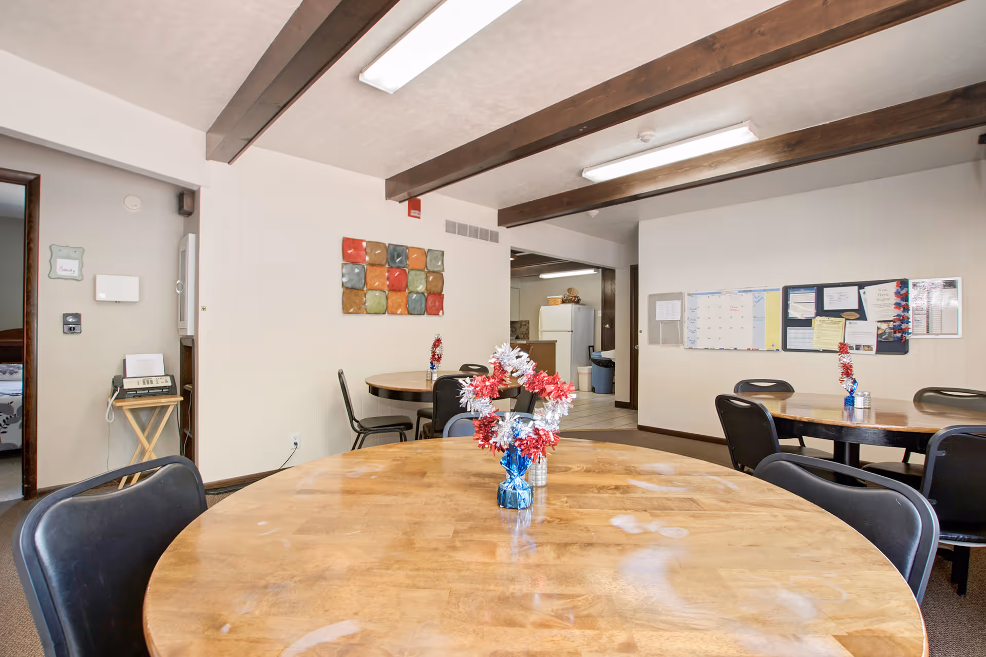 Interior view of a common dining area in an assisted living facility with round wooden tables and black chairs. Each table has a small patriotic-themed centerpiece with red, white, and blue decorations. The room has white walls, exposed wooden ceiling beams, and a bulletin board with various notices on one wall. In the background, there is a kitchen area with a refrigerator and a doorway leading to another room.