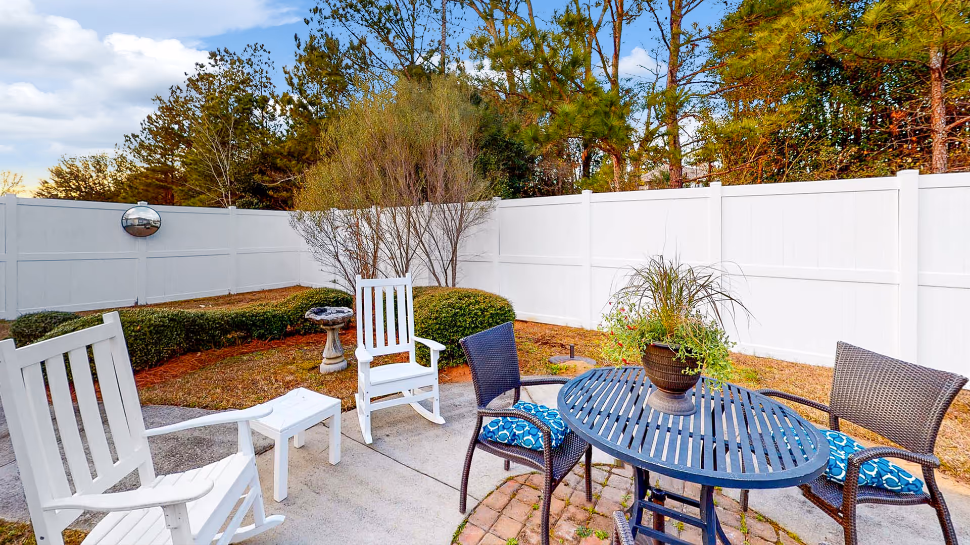 Fenced outdoor patio area with a round metal table, chairs with blue cushions, white rocking chairs, and a potted plant.