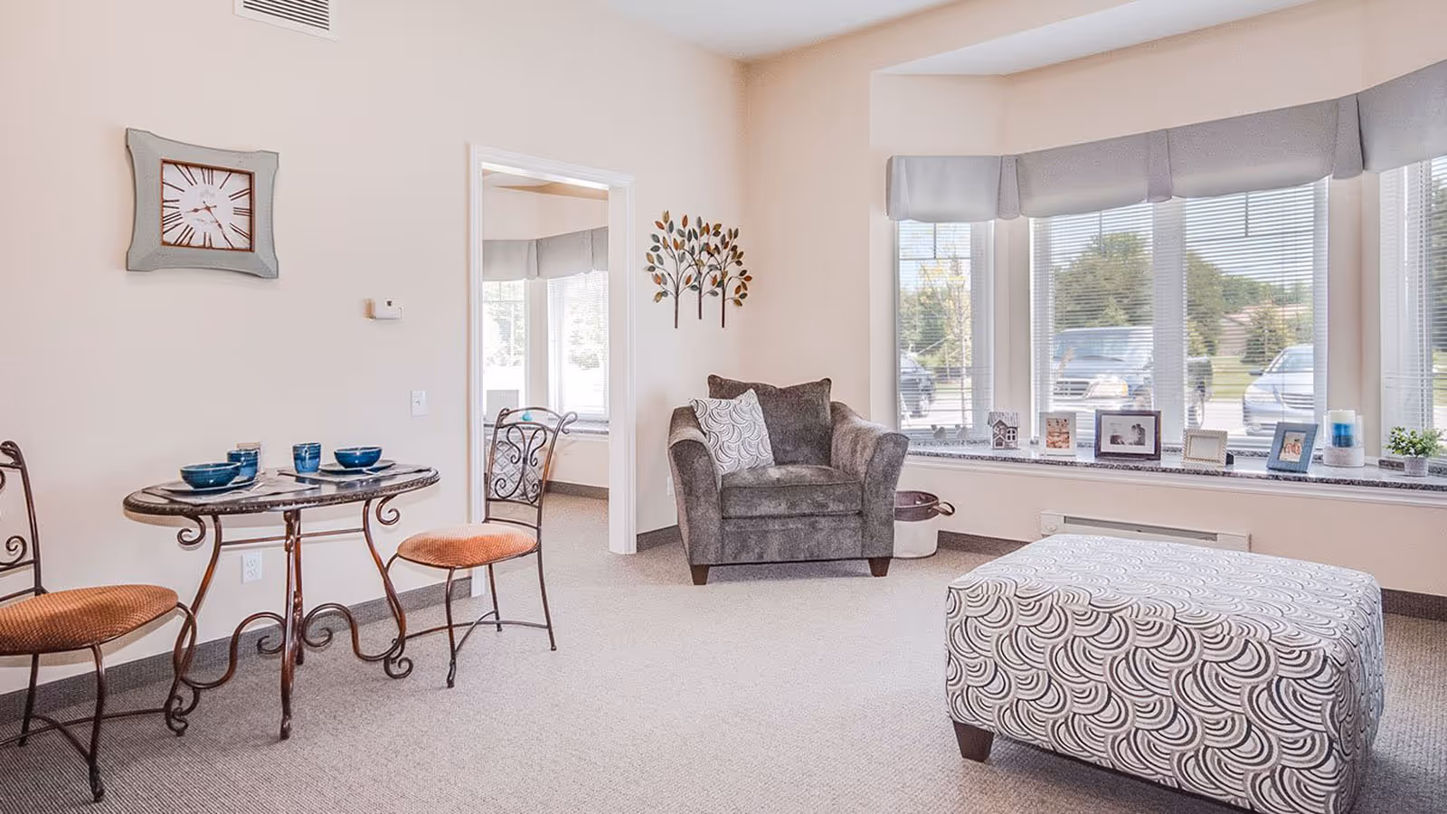 A bright and cozy living area with a small round table set with blue bowls and cups, two chairs with orange cushions, a gray armchair with decorative pillows, and a patterned ottoman. Large windows with blinds and a valance let in natural light, and a wall clock and decorative metal wall art are visible.