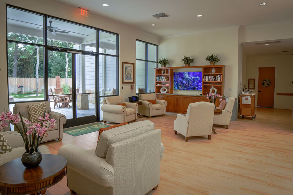 A bright and spacious living room area in a senior living facility with several beige upholstered armchairs arranged around a wooden coffee table with a vase of pink flowers. Large glass doors and windows provide a view of an outdoor patio with chairs and greenery. A wooden entertainment center with shelves, plants, and a large screen displaying an aquarium scene is against one wall.