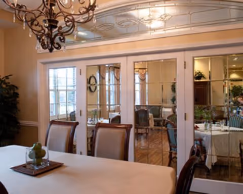 A dining room with a long table covered with a white tablecloth, surrounded by chairs. A decorative chandelier hangs above the table. In the background, there are glass-paneled double doors leading to another room with additional dining tables and chairs. The room has warm lighting and elegant decor.