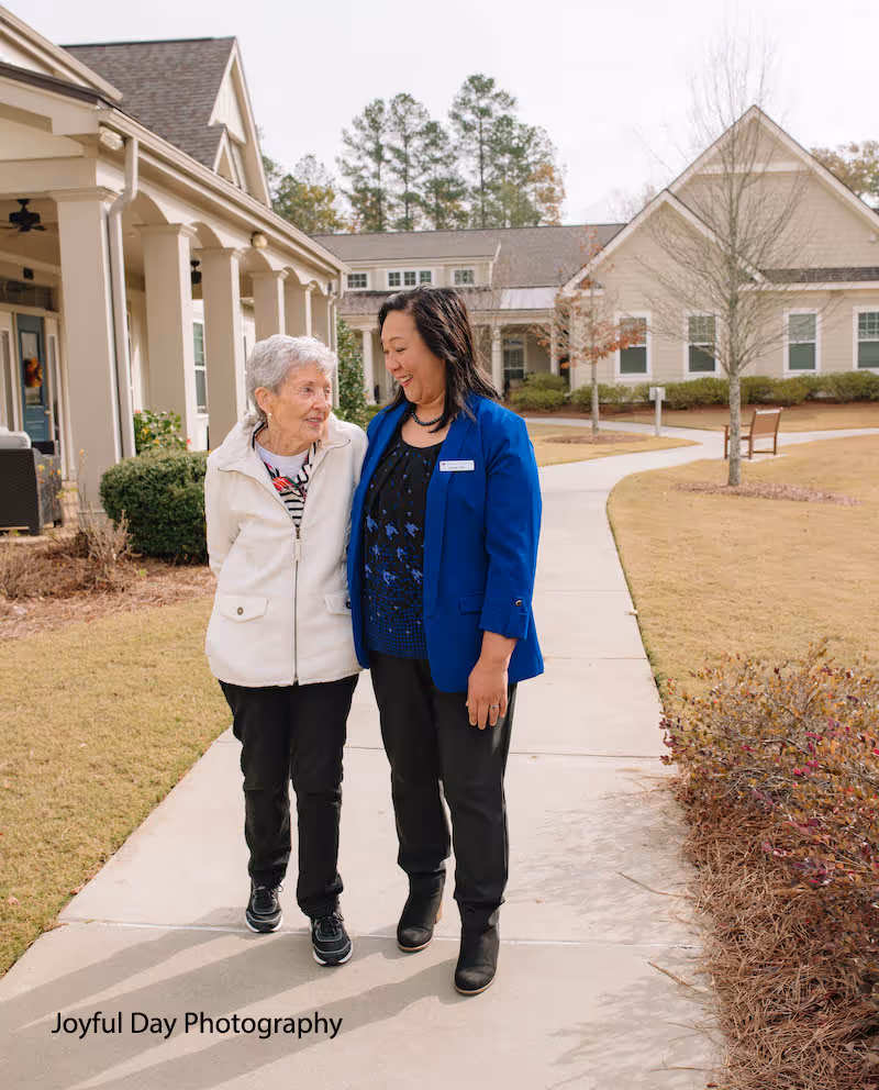An elderly woman and a caregiver walk arm in arm along a sidewalk outside a senior living community. They are smiling and looking at each other warmly. The background shows beige buildings, some bushes, and trees with no leaves.