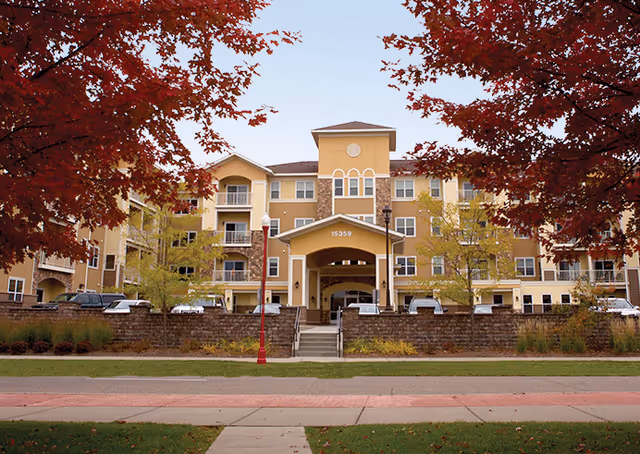 Front exterior view of a multi-story senior living facility building with beige and yellow walls, balconies, and a covered entrance. The building is framed by red autumn trees and a sidewalk in the foreground.