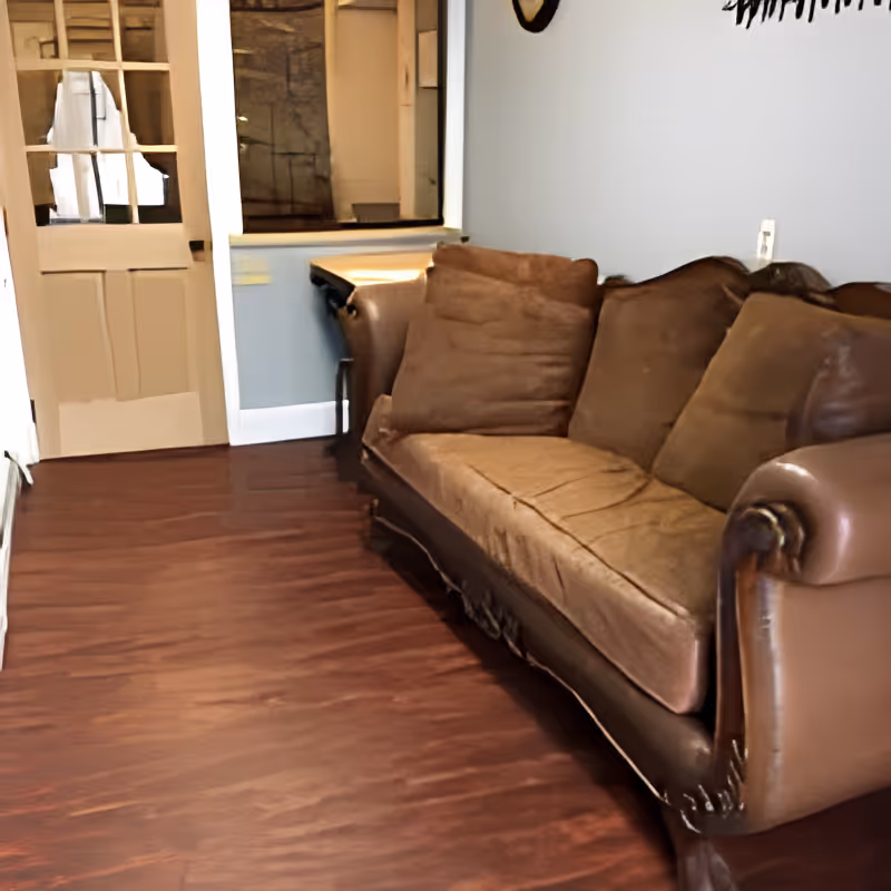 A brown leather and fabric sofa with several cushions is placed against a light blue wall in a room with dark wood flooring. To the left, there is a door with glass panes and a small window opening above a corner table.