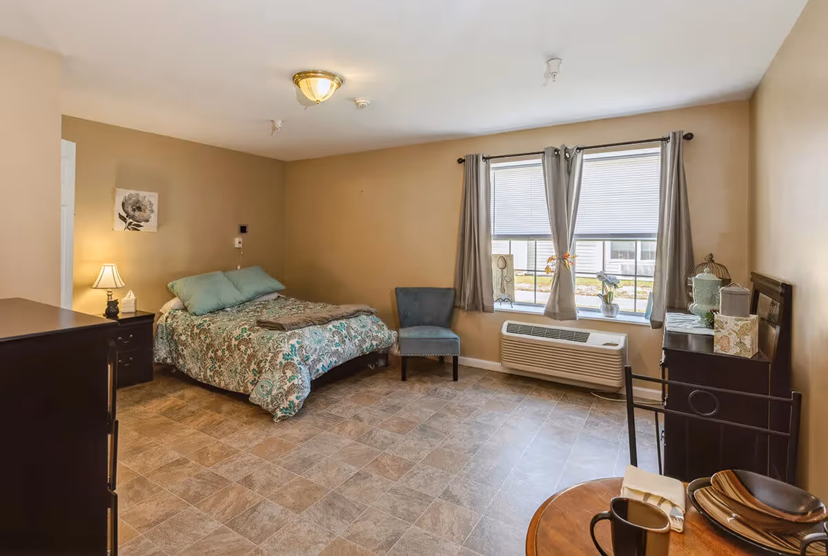 A furnished bedroom with a bed, bedside table and lamp, dresser, chair, window with curtains, and a small table with dishes.