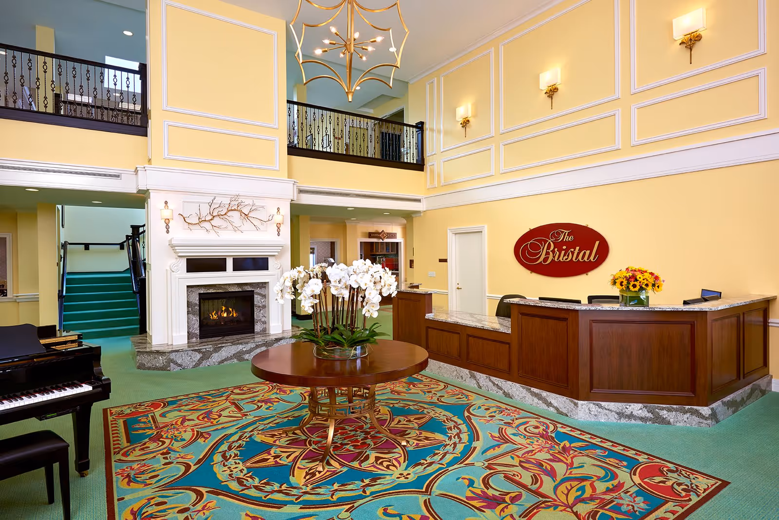 A bright and elegant assisted living facility lobby with yellow walls and white trim, a large colorful patterned rug, a round wooden table with a white orchid centerpiece, a grand piano, a fireplace with a decorative branch above it, a wooden reception desk with a flower arrangement, and a sign that reads 'The Bristal'.