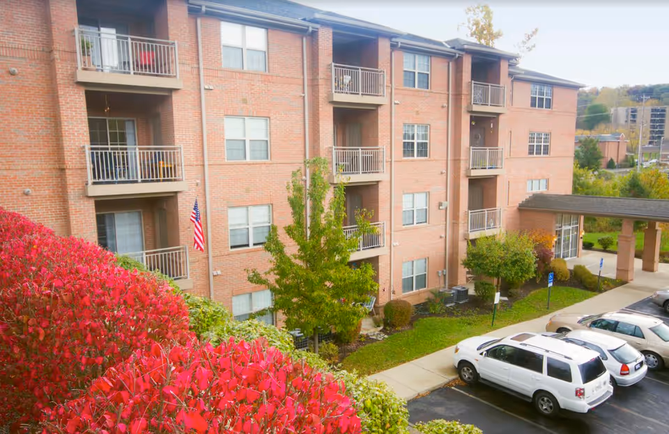 Exterior view of a multi-story brick residential building with balconies, surrounded by green trees and red bushes. Several cars are parked in the parking lot in front of the building, and an American flag is displayed near one of the balconies.