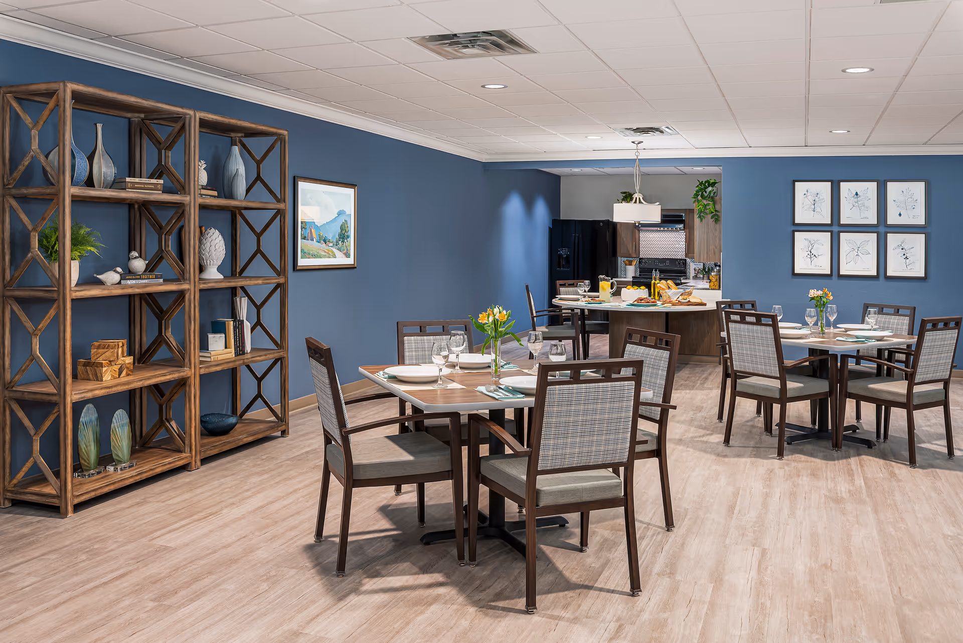 A dining room with several wooden tables and chairs set with plates, glasses, and cutlery. The room has blue walls, light wood flooring, and a wooden shelving unit with decorative items on the left. In the background, there is a kitchen area with a black refrigerator and a countertop with food and drinks. Artwork and framed pictures decorate the walls.