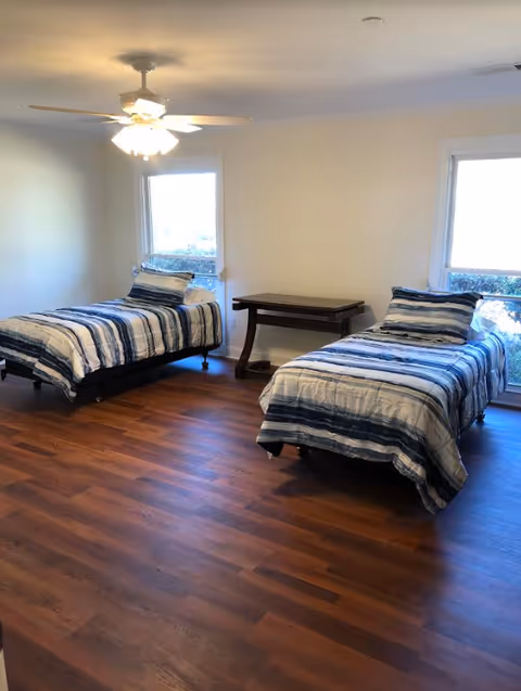 A bedroom with two single beds, each with blue and white striped bedding and pillows. There is a wooden floor, a ceiling fan with lights, two windows letting in natural light, and a small wooden table between the beds.