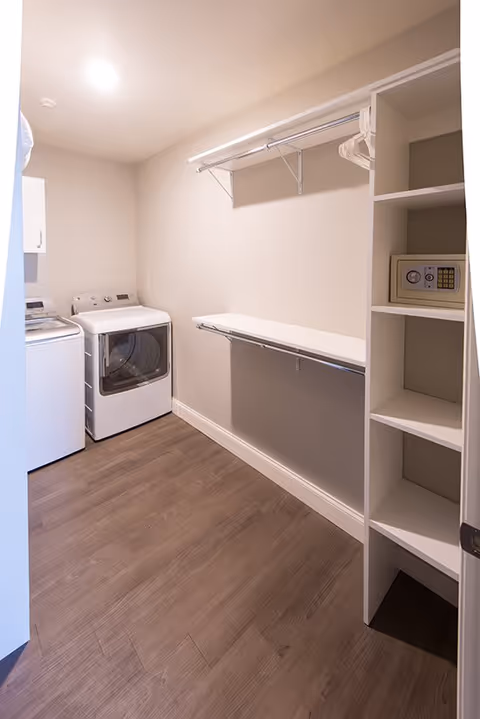 A laundry room with a washing machine and dryer on the left side, a white shelf with hangers above a folding counter on the right wall, and a shelving unit with a small safe on the right side. The room has wood flooring and beige walls with a ceiling light.
