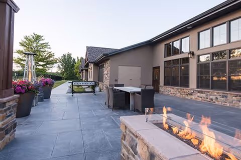Outdoor patio area at Ballard Glenn featuring a modern fire pit with flames, a dining table with chairs, large planters with flowers, and a building with large windows in the background under a clear sky.