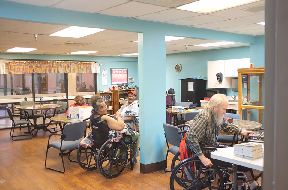 A common area in an assisted living facility with several elderly residents sitting at tables and in wheelchairs. The room has light blue walls, wooden floors, and large windows with curtains. There are tables and chairs arranged throughout the space, and shelves with books and decorations in the background.