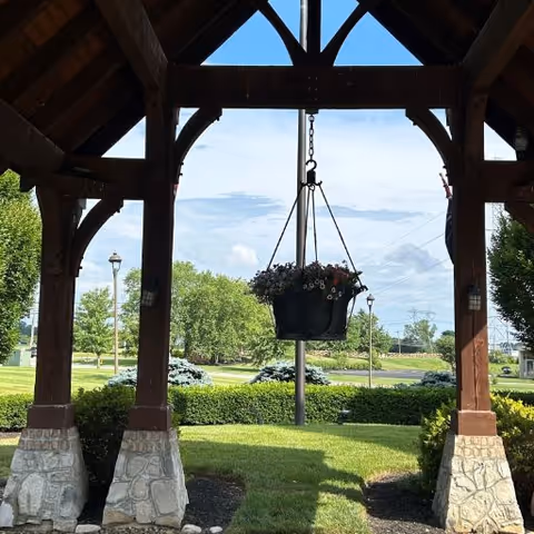 View from under a wooden gazebo with stone pillars, looking out onto a landscaped garden area with green grass, bushes, trees, and a clear blue sky with some clouds. A hanging flower pot with blooming flowers is suspended from the center of the gazebo roof.