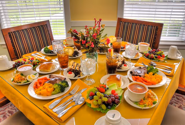 A dining table set with a yellow tablecloth and four place settings, each with a plate of food including vegetables, fruit, rolls, and desserts. There are glasses of iced tea and water, coffee cups, and a centerpiece with colorful flowers. The table is surrounded by wooden chairs with striped cushions, and windows with blinds are in the background.