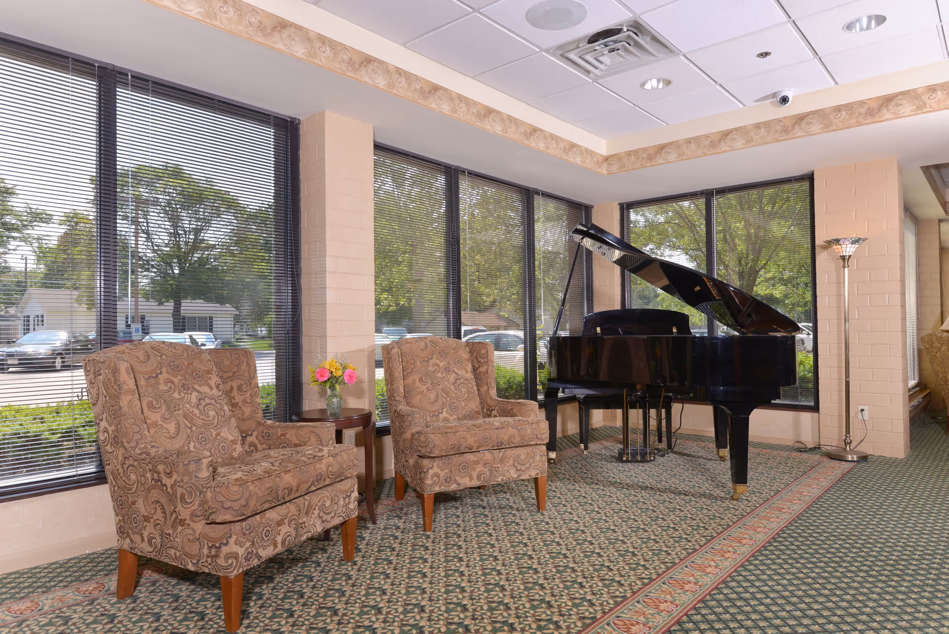 A cozy sitting area with two patterned armchairs and a small round table with a vase of flowers between them, next to a black grand piano. Large windows with blinds let in natural light, showing trees and parked cars outside. The room has a patterned carpet and beige walls with decorative trim near the ceiling.