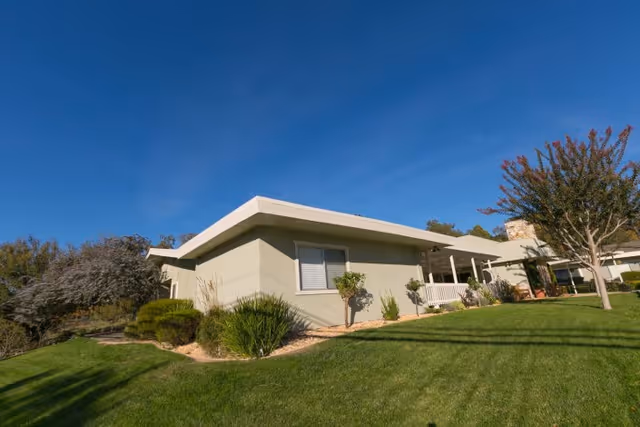 Exterior view of a single-story building with light-colored walls and a flat roof, surrounded by a well-maintained green lawn, shrubs, and trees under a clear blue sky.