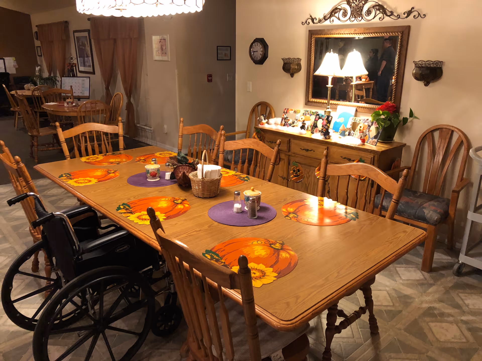 Communal dining table with pumpkin-themed placemats, wooden chairs and a wheelchair in a warmly decorated dining room.