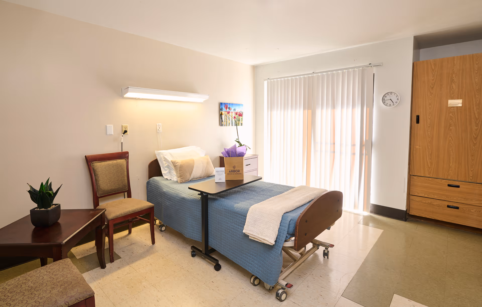 A tidy nursing facility bedroom with a single hospital bed, overbed table, chairs, a plant, and vertical blinds letting in sunlight.