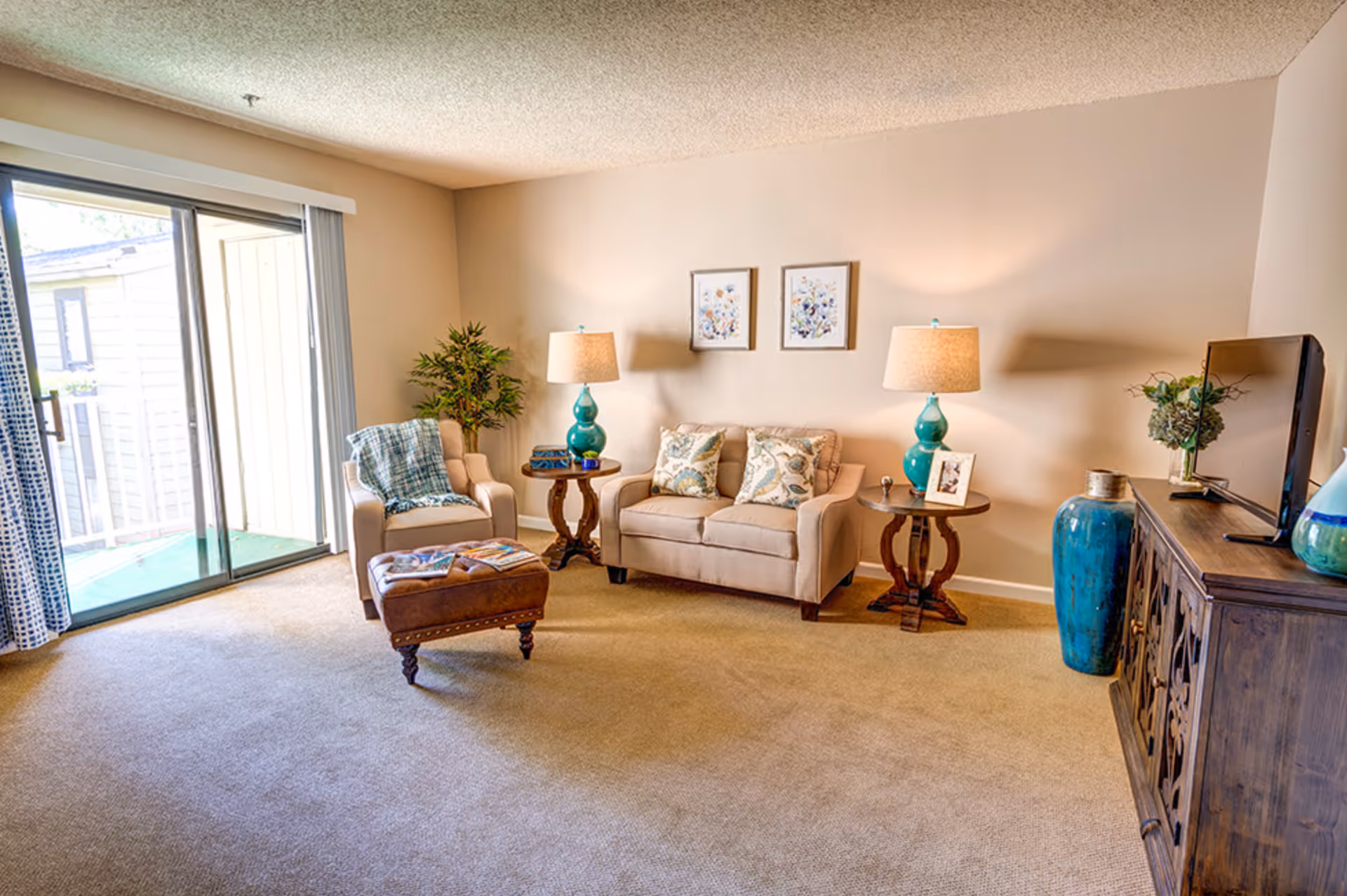 A cozy living room with beige carpet and walls, featuring a beige loveseat with patterned cushions, a matching armchair with a blue throw blanket, two wooden side tables with turquoise lamps, a brown leather ottoman with magazines on top, a wooden cabinet with a TV and decorative items, and a sliding glass door leading to an outdoor balcony.