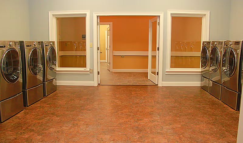 A spacious laundry room with front-loading washers lined along both walls and a doorway leading to a hallway.