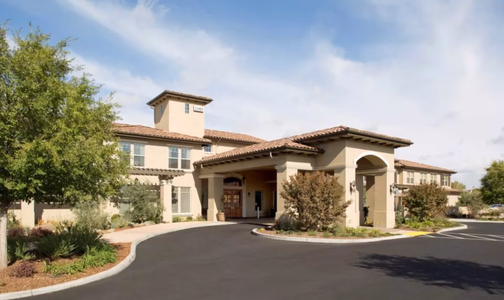 Front exterior of a beige Mediterranean-style senior living building with a covered entrance, driveway, and landscaped grounds.