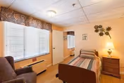A small, cozy bedroom with a single bed covered in a striped blanket, a wooden nightstand with a lamp, a cushioned armchair, and a window with blinds and a decorative valance. The walls are painted a soft peach color and decorated with a metal wall art piece and a small framed picture.