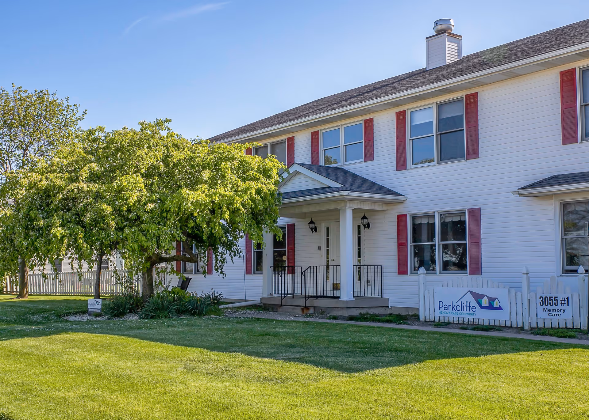 Exterior view of a two-story white building with red window shutters and a small covered entrance porch. There is a green lawn and a leafy tree in front of the building. A white fence with a sign reading 'Parkcliffe Memory Care Community' and '3055 #1 Memory Care' is visible near the entrance.