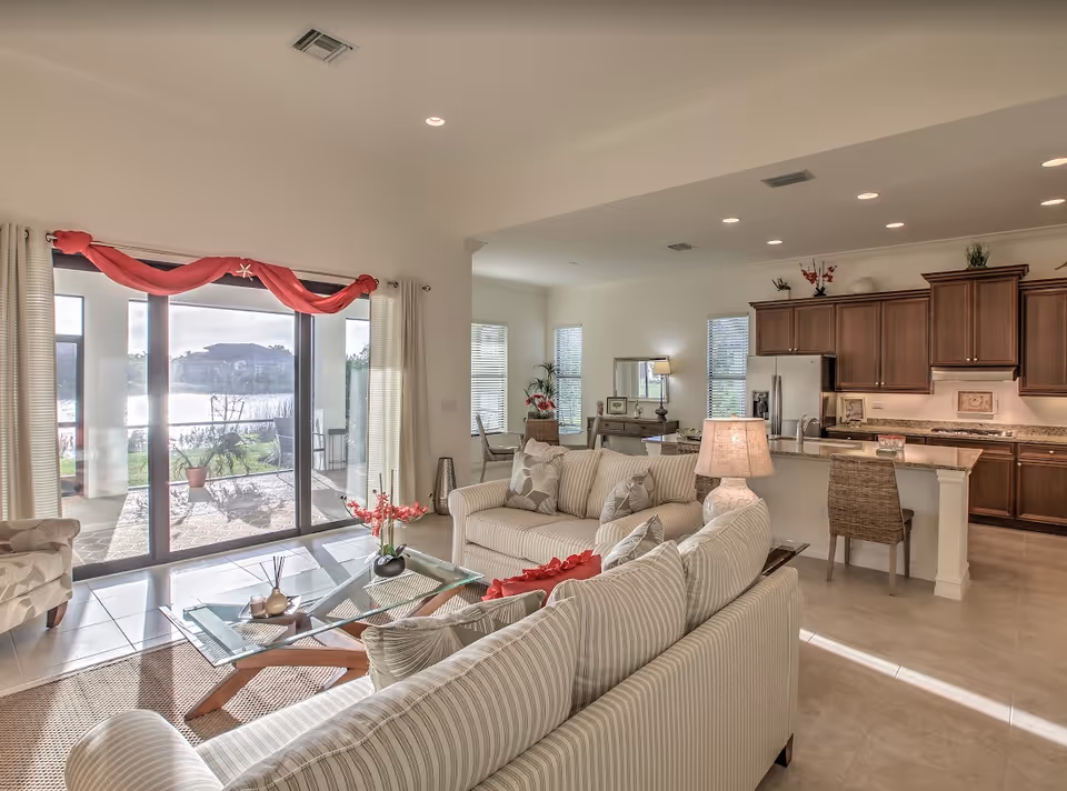 Bright open living room and kitchen with striped sofas, glass coffee table, and sliding glass doors overlooking a lakeside patio.