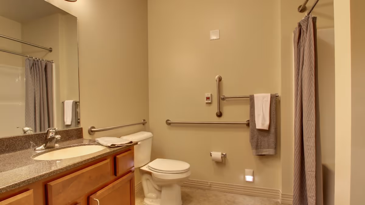 A clean and accessible bathroom with a toilet, a sink with a granite countertop, a large mirror, grab bars on the walls, a towel rack with towels, and a shower with a curtain.