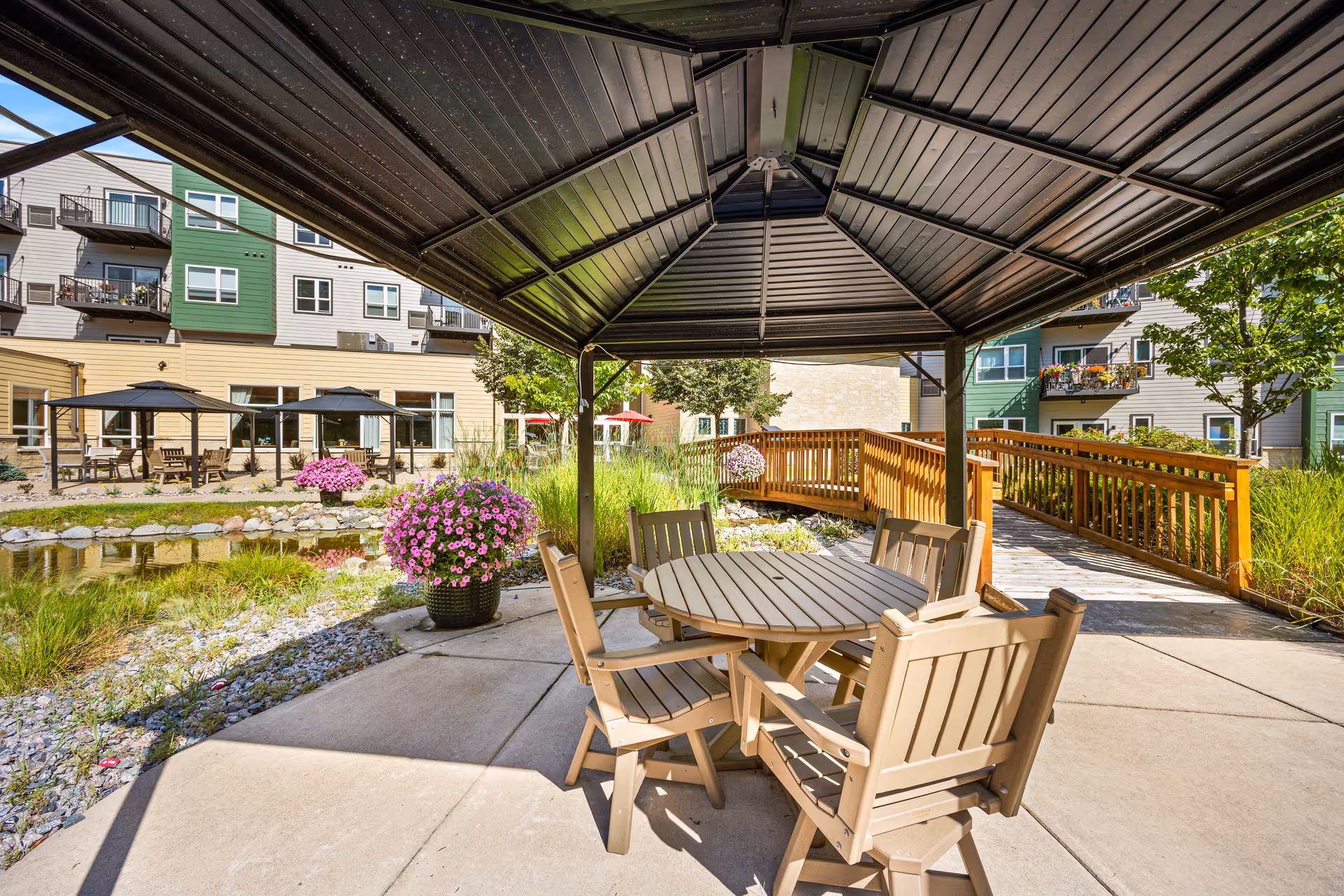 Outdoor seating area at Woodland Hill featuring a round table with four chairs under a metal gazebo. Surrounding the area are landscaped plants, a small pond with rocks, wooden walkways, and multi-story residential buildings with balconies in the background.