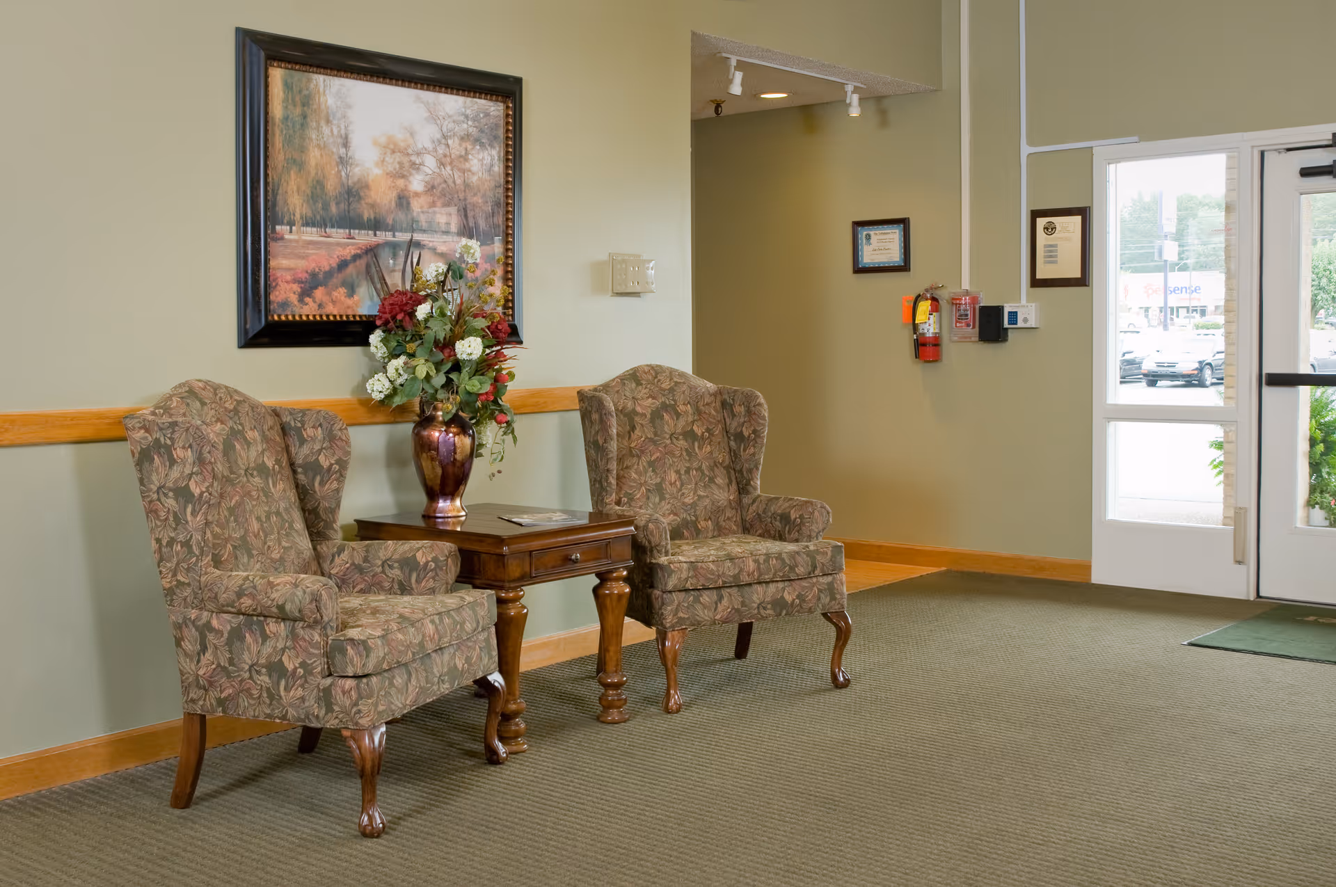 Lobby seating area with two patterned armchairs flanking a wooden side table holding a flower vase near a glass entrance door.