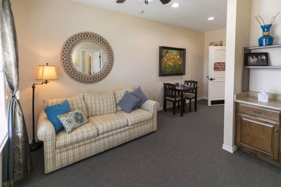 A cozy living room area with a plaid yellow sofa adorned with blue and patterned cushions. A floor lamp stands next to the sofa near a window with curtains. Above the sofa is a large decorative round mirror. In the background, there is a small dining table with four chairs and a framed painting on the wall. To the right, there is a wooden cabinet with a blue vase and a framed photo on the shelf. The room has beige walls and carpeted flooring.