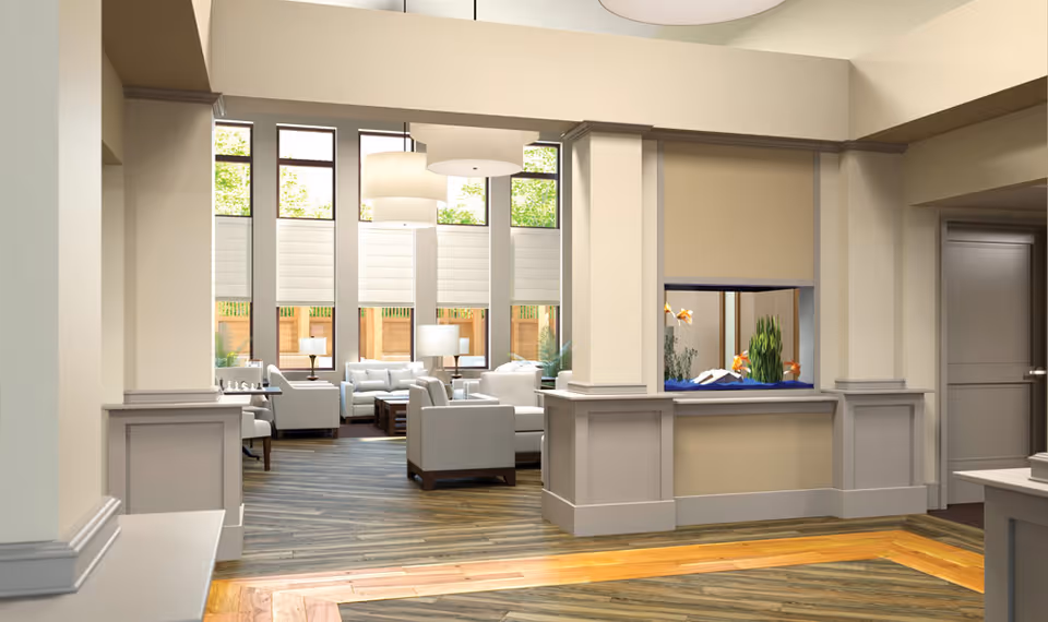 A bright and spacious living room area with large windows letting in natural light. The room features modern white armchairs and sofas arranged around wooden coffee tables. There are two large pendant lights hanging from the ceiling and table lamps near the seating. A built-in aquarium with fish is set into a wall partition. The flooring is a mix of wood patterns, and the walls are painted in neutral tones.