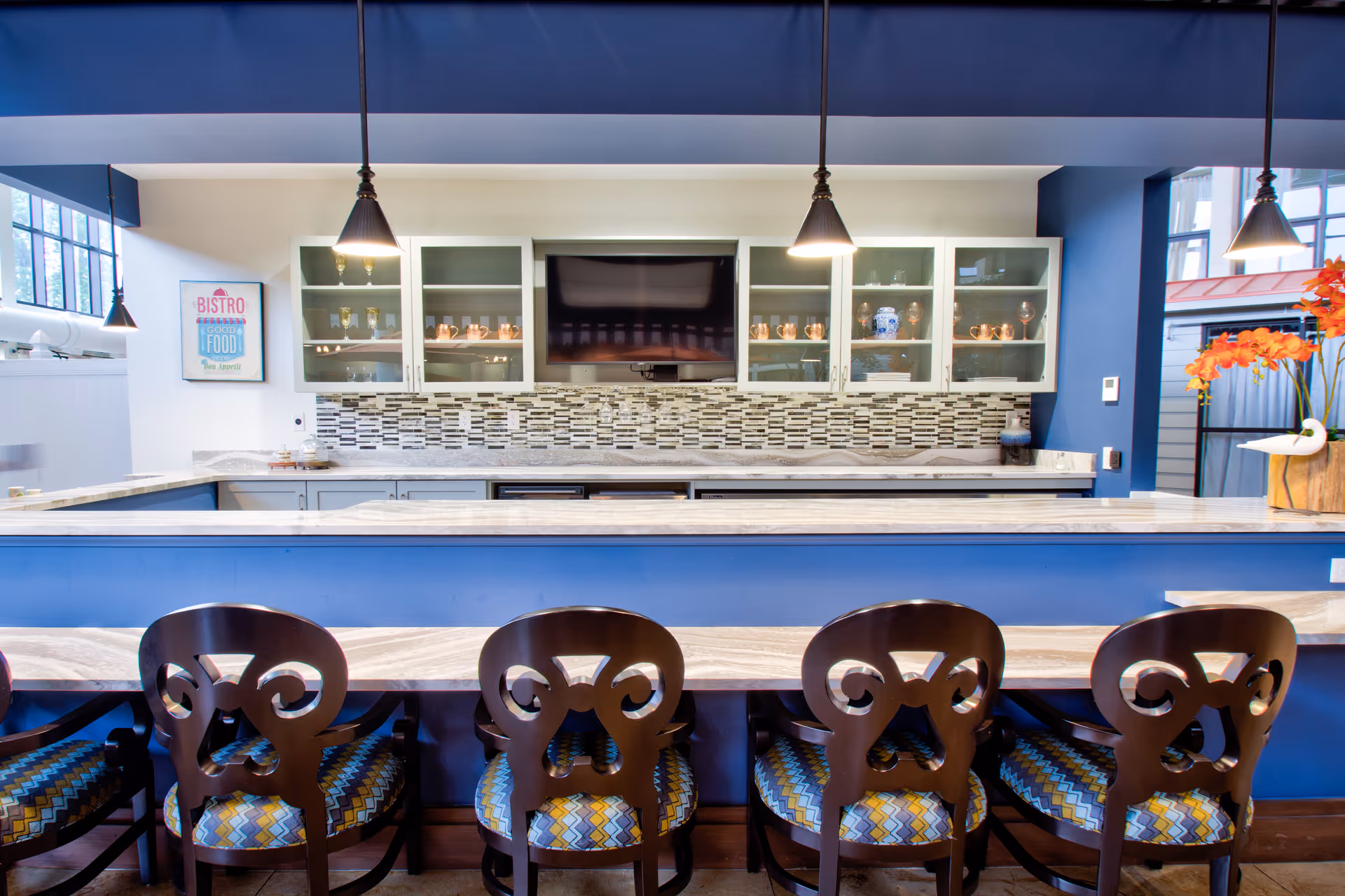 Interior view of a modern kitchen area with a marble countertop bar and five wooden chairs with patterned cushions. Above the counter are three pendant lights. Behind the counter are glass-front cabinets, a mounted flat-screen TV, and a tiled backsplash. A framed sign on the left wall reads 'Bistro Fresh Food'. On the right side, there is a vase with orange flowers.
