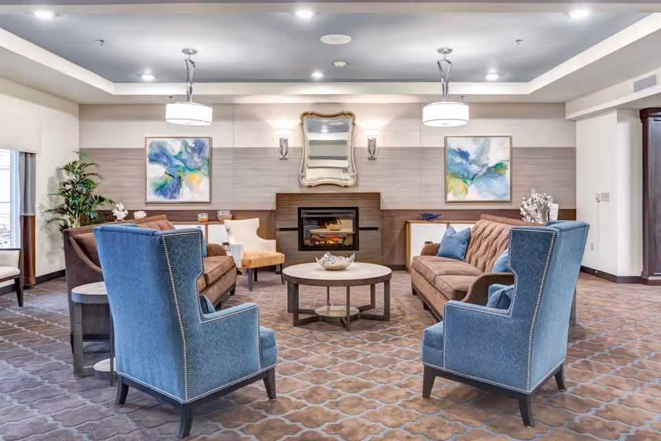 A spacious and well-lit living room area featuring two blue upholstered armchairs facing two brown sofas around a round coffee table. The room has a patterned carpet, a fireplace with a decorative mirror above it, two abstract paintings on the wall, and modern ceiling lights. There are also plants and decorative items on side tables and cabinets.
