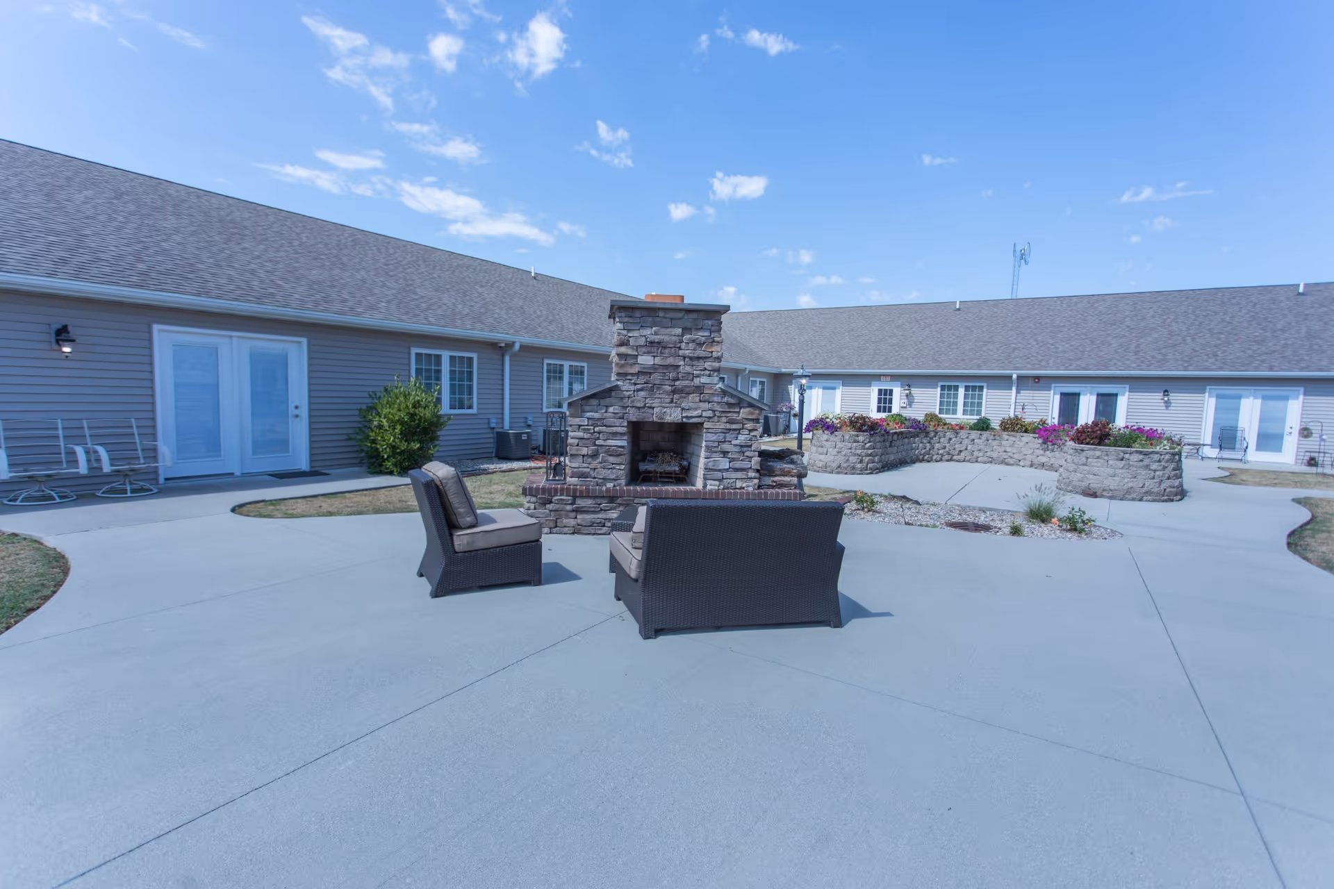 Outdoor courtyard area of a senior living facility with a stone fireplace in the center, surrounded by cushioned wicker chairs. The courtyard is paved with concrete and has flower beds with colorful flowers along the building walls. The building has beige siding, multiple windows, and several doors leading to the courtyard. The sky is clear with a few clouds.