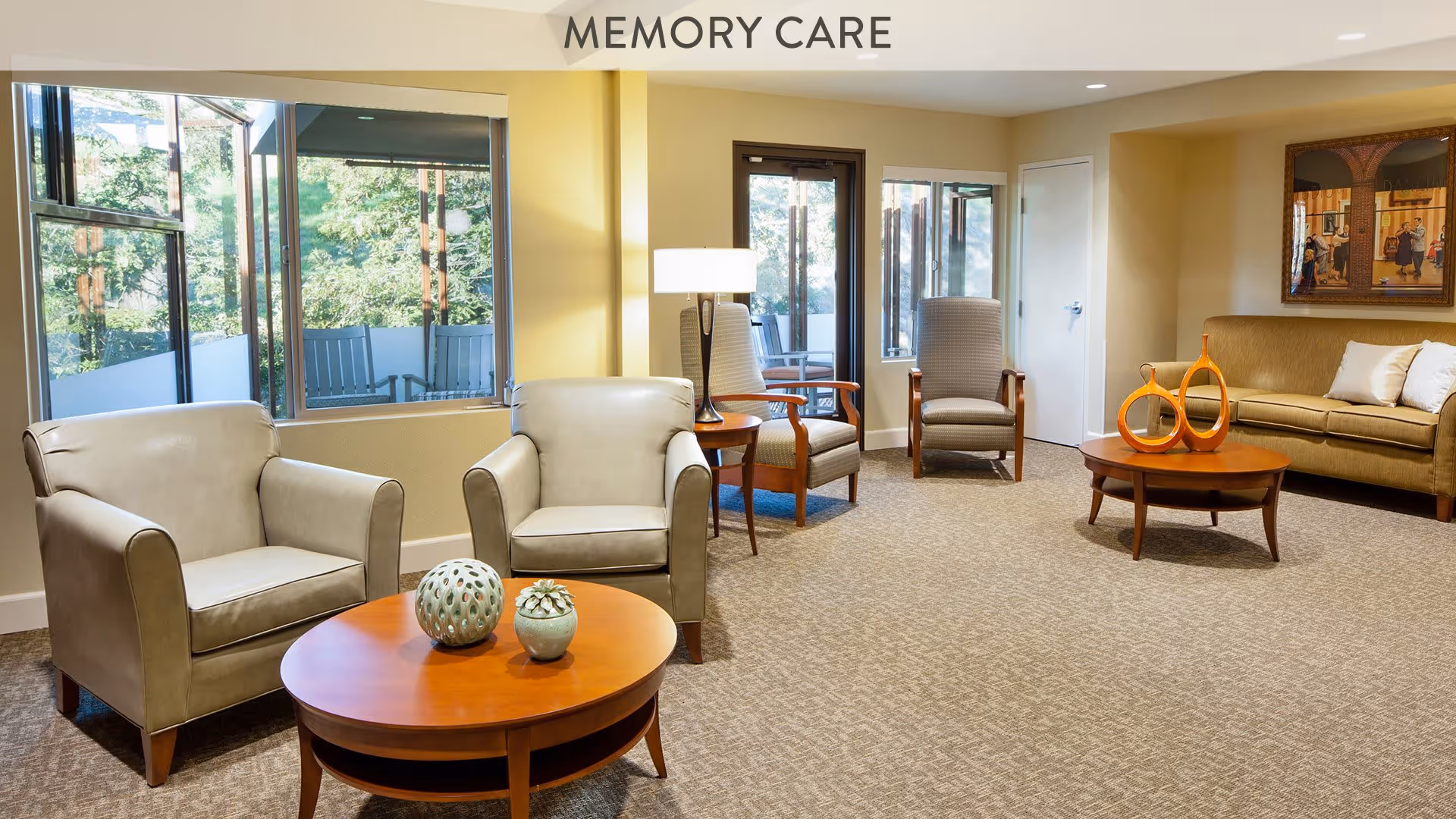 A well-lit memory care lounge area with beige walls and carpet. The room features two beige armchairs around a wooden coffee table with decorative items, two wooden chairs with cushions near a window, and a beige sofa with pillows. Large windows provide a view of greenery outside, and a floor lamp is placed between the armchairs and chairs. A framed artwork hangs on the wall above the sofa.