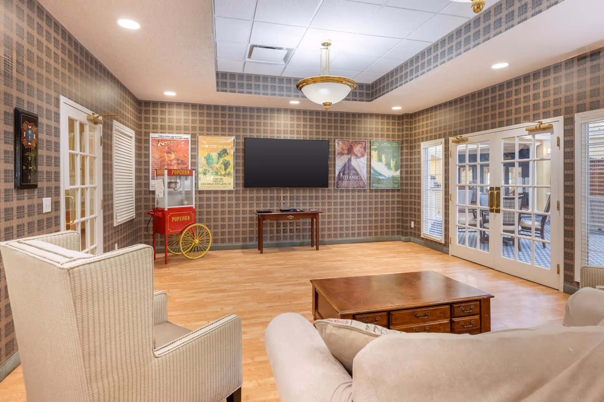 A cozy living room area with patterned wallpaper, a large flat-screen TV mounted on the wall, a vintage-style popcorn machine, and framed movie posters. The room features comfortable seating including an armchair and a sofa, a wooden coffee table, and French doors leading to another room with additional seating.
