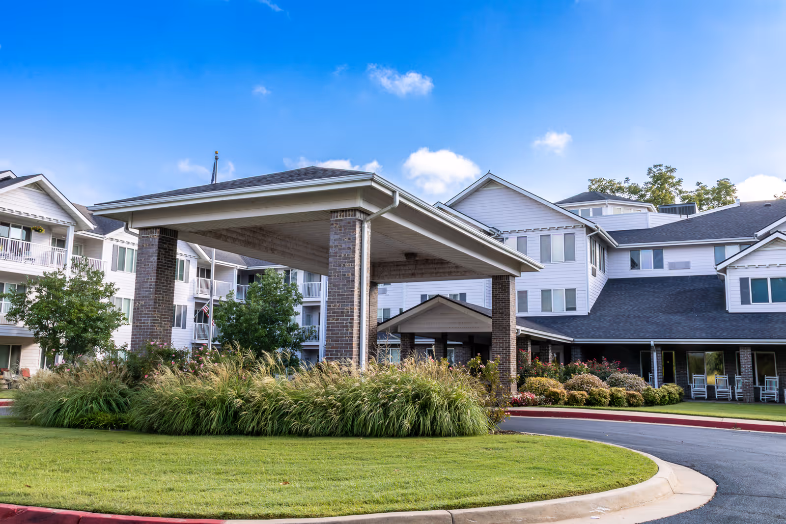 Exterior view of Asher Point Independent Living of Tulsa, showing a large covered entrance with brick pillars, surrounded by well-maintained landscaping including green grass, bushes, and trees under a blue sky with some clouds.