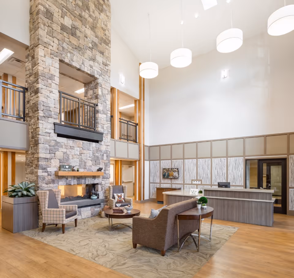 A spacious and well-lit senior living facility lobby with a tall stone fireplace as the focal point. The room features a seating area with a brown sofa, two patterned armchairs, a round coffee table, and a side table with plants. Behind the seating area is a reception desk with a welcoming sign and a small potted plant. The walls are decorated with wood paneling and a mounted television. Large pendant lights hang from the high ceiling, and there is a second-floor balcony visible above the fireplace.
