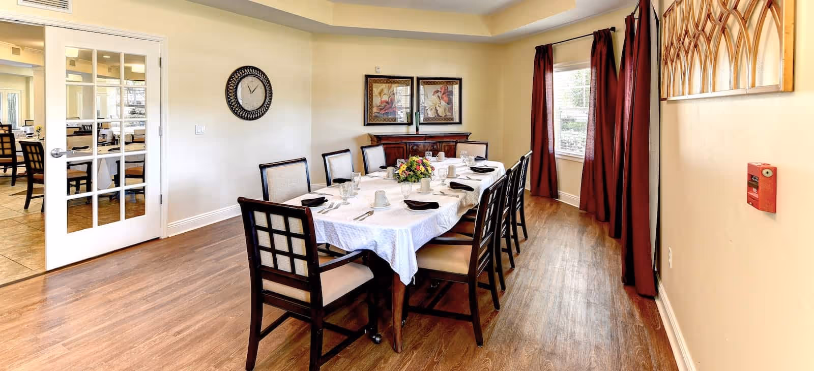 A dining room with a long rectangular table covered with a white tablecloth, set with plates, cups, glasses, and black napkins. The table is surrounded by eight wooden chairs with cushioned seats. The room has wooden flooring, beige walls, a wall clock, two framed pictures, and red curtains on the windows. A glass door leads to another dining area in the background.