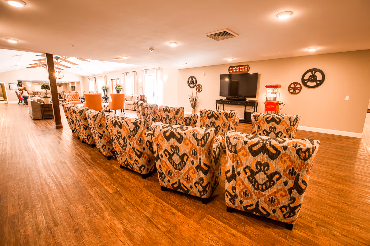 A spacious senior living facility common area with a row of patterned armchairs facing a large flat-screen TV mounted on the wall. The room has wooden flooring, beige walls, and several decorative film reels on the wall. There is a red popcorn machine next to the TV, and additional seating with sofas and chairs is visible in the background near large windows letting in natural light.