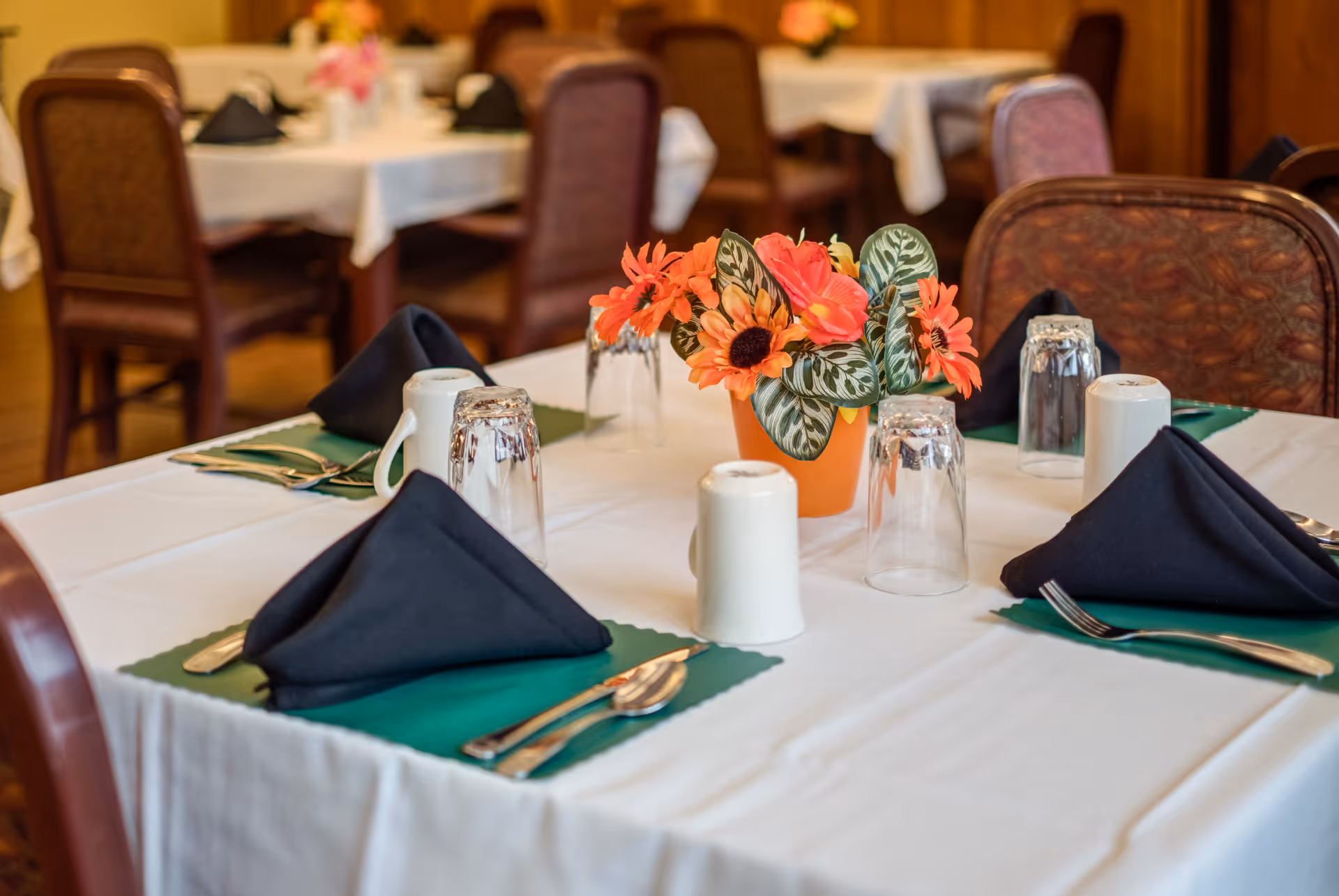 A dining table set with white tablecloth, green placemats, black folded napkins, silverware, upside-down drinking glasses, white coffee mugs, and a small orange pot with artificial orange and pink flowers in the center. Several similar tables and chairs are visible in the background in a warmly lit dining room.